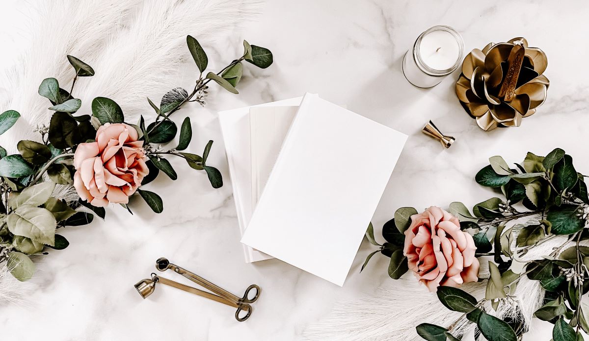 three white books surrounded by pink roses, greenery, a candle, wick trimmer, and snuffer on a marble background